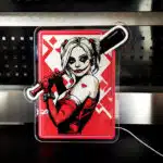 Podświetlony neon LED przedstawiający postać kobiety z kijem bejsbolowym na tle czerwonych wzorów.